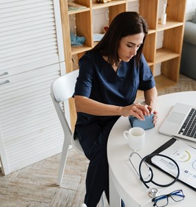 Kvinna i sjukvårdskläder sitter vid dator och öppnar sin plånbok.